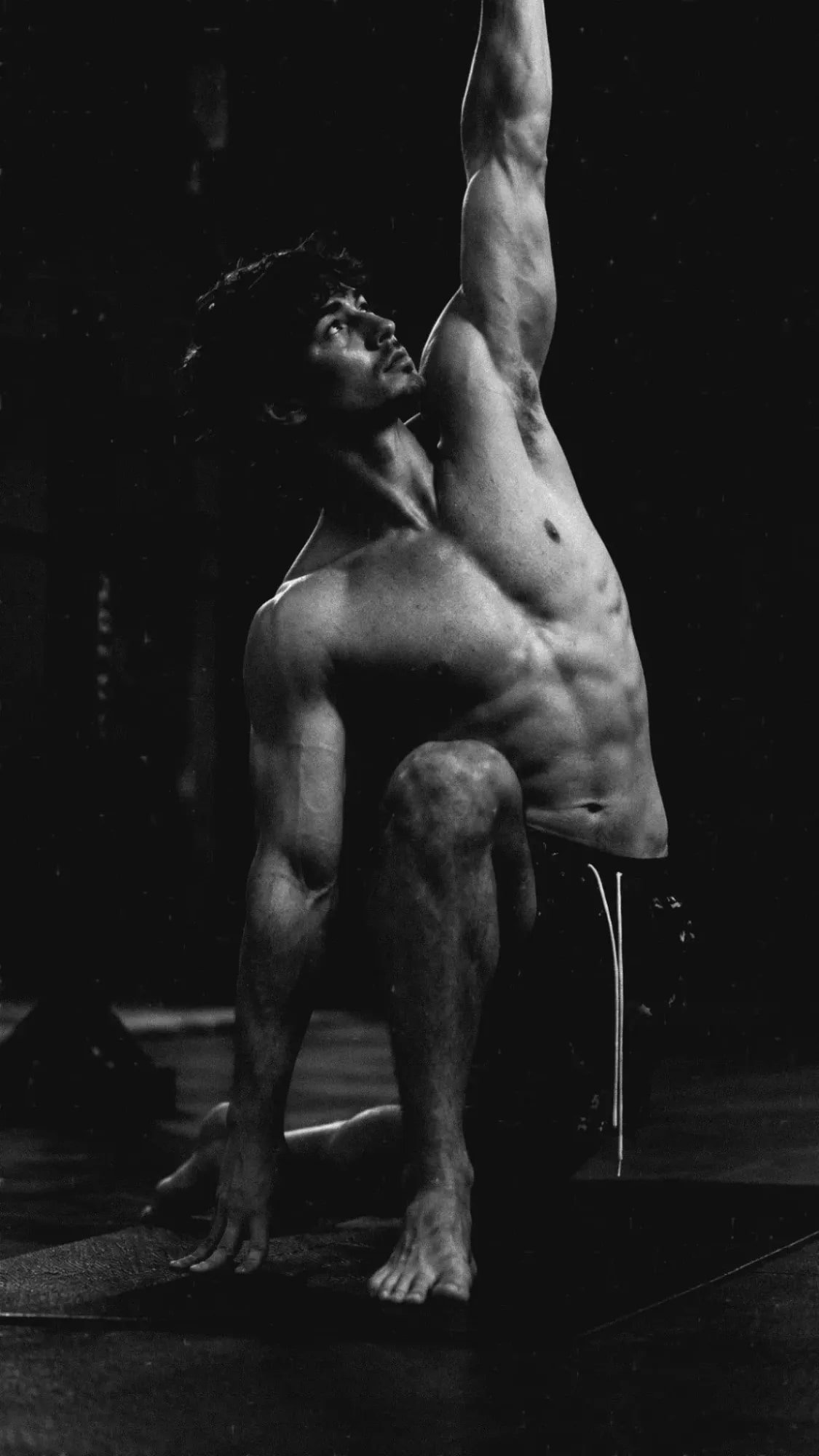 Andrew Valentine kneeling on a gym floor, one arm reaching straight overhead, looking up along it, lit from the side against a dark ground.
