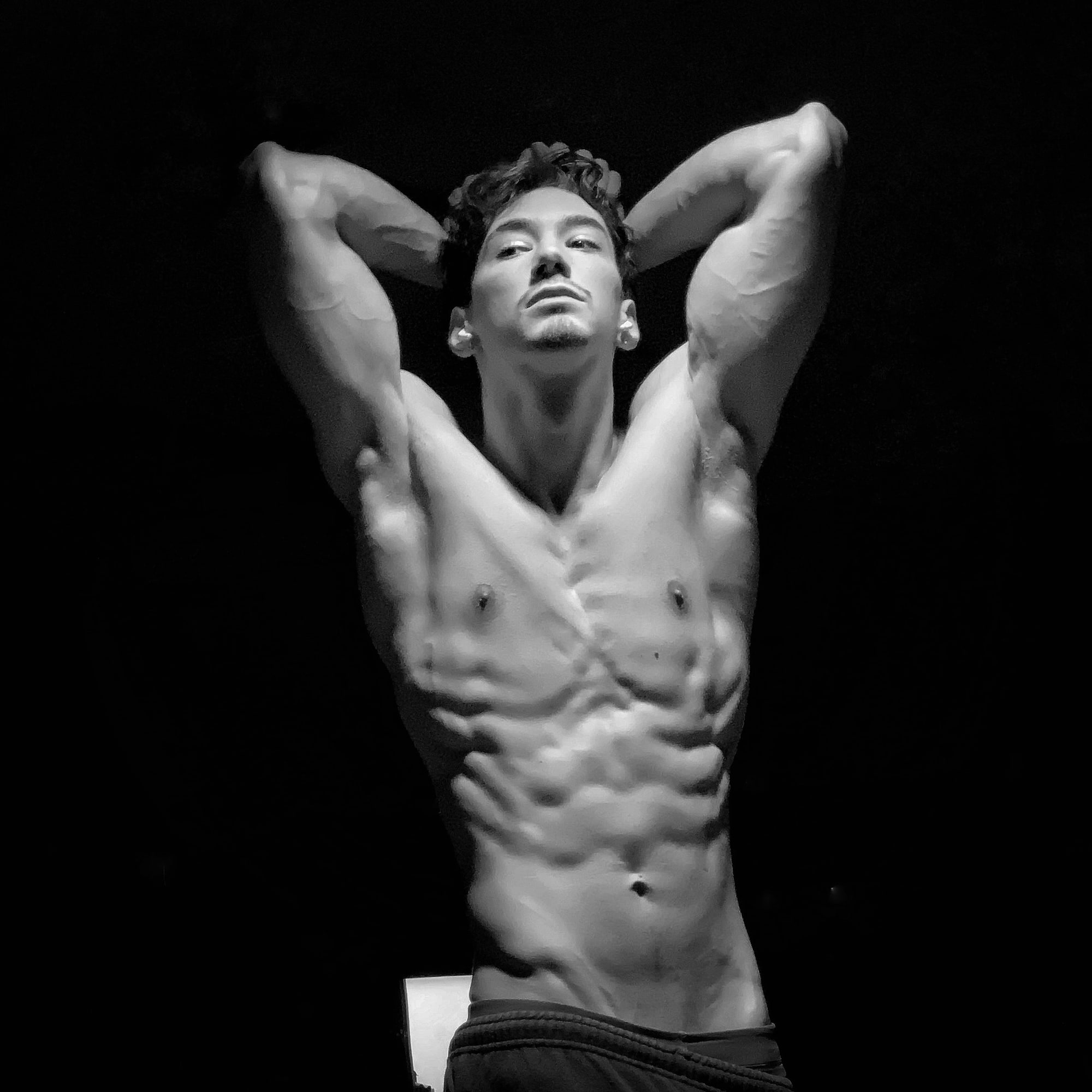 Andrew Valentine, arms behind his head, lit from above against a dark ground.