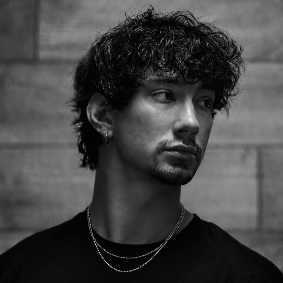Andrew Valentine, head and shoulders, looking off to the side against a stone wall. Curly dark hair, a small hoop earring, two thin chains.