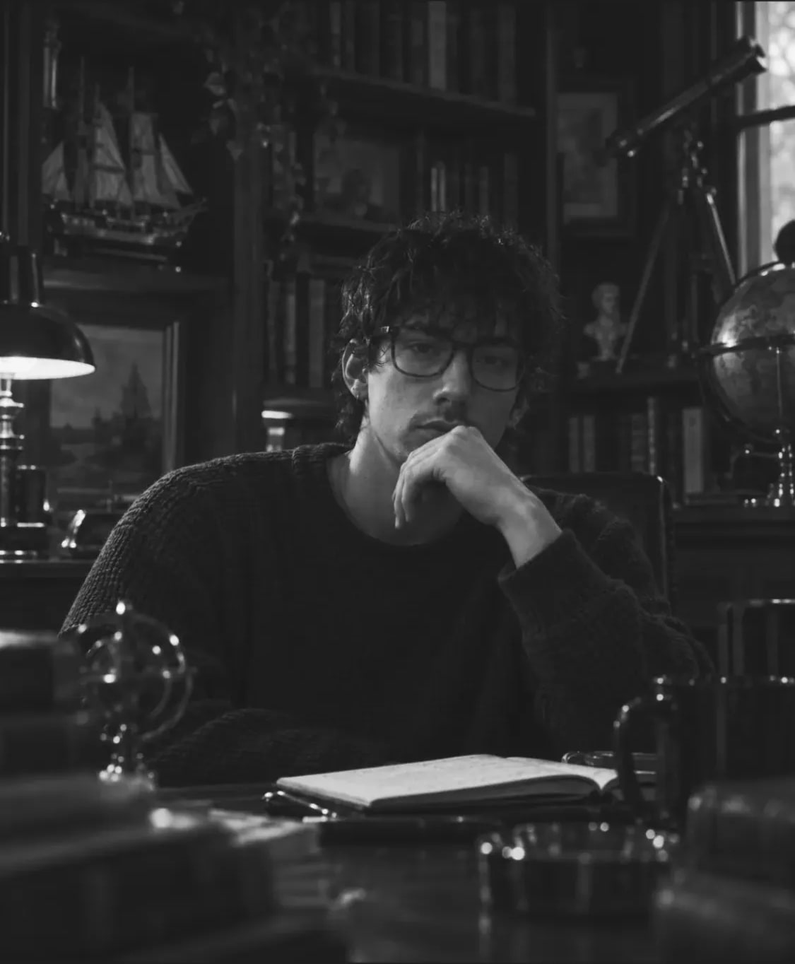 Andrew Valentine in glasses and a dark sweater at a desk in a book-lined study, chin on his hand, an open notebook in front of him.